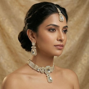 Op de afbeelding: Een goudkleurige sieradenset met een ketting, oorbellen en een hoofdband. De ketting heeft een rechthoekige centrale hanger en is versierd met parels. De oorbellen en hoofdband weerspiegelen het hangerontwerp, allemaal gezet met heldere, sprankelende stenen.