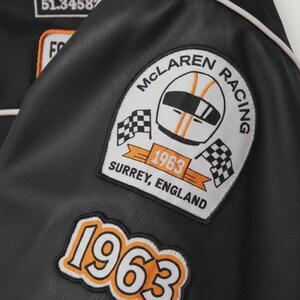 May include: Black leather jacket with white piping and patches. Patches include "McLaren Racing" logo with a helmet and checkered flags, and the year "1963". The jacket also has the text "Surrey, England".