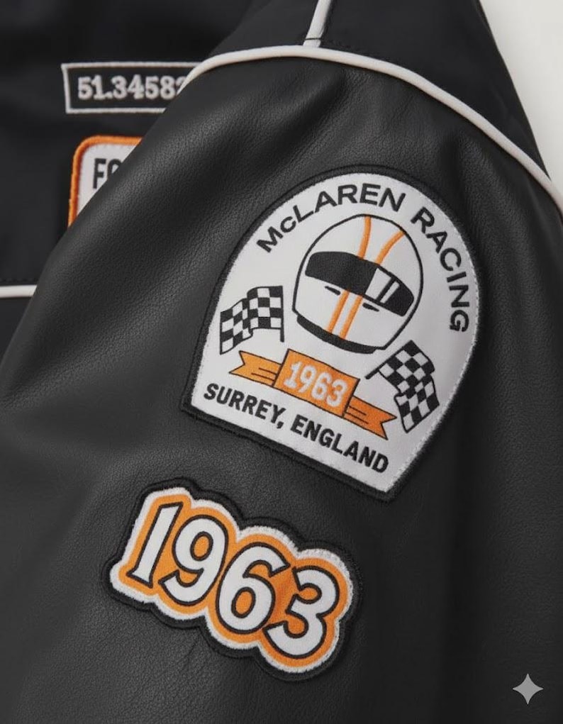May include: Black leather jacket with white piping and patches. Patches include "McLaren Racing" logo with a helmet and checkered flags, and the year "1963". The jacket also has the text "Surrey, England".