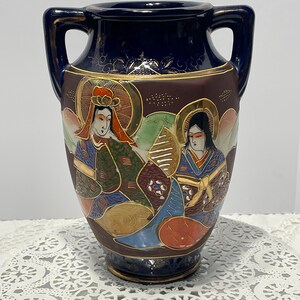 May include: A dark blue ceramic vase with two handles, featuring a hand-painted scene of two figures in traditional attire with gold accents. The design includes intricate patterns, a dark brown central band, and gold-colored decorative details.