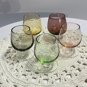 May include: Five colorful stemmed glasses in shades of green, yellow, pink, and orange are arranged on a cream-colored, crocheted doily. The glasses have rounded bowls and clear stems.