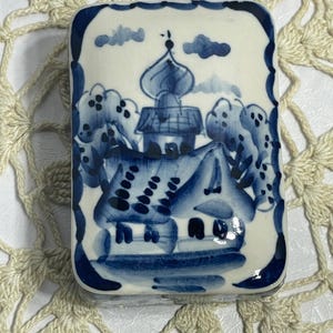 May include: Rectangular ceramic trinket box with a blue and white hand-painted design. The box features a detailed illustration of a building with a domed roof, trees, and clouds. The edges of the box are outlined in blue.