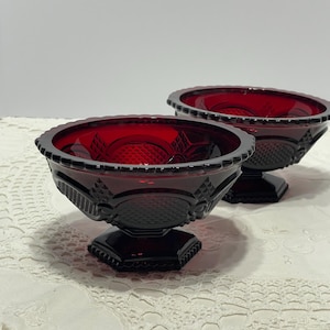 May include: Two matching ruby red glass bowls featuring a pressed pattern with diamond and circular motifs. Each bowl has a scalloped rim, a wide body, and sits on a hexagonal pedestal base. The glass is translucent and deep red in color.