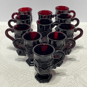 May include: A set of ten ruby red glass mugs arranged in a triangular formation. Each mug features a textured diamond and circle pattern on the exterior, a curved handle, and a hexagonal base. The glass is translucent, showing a deep red color.