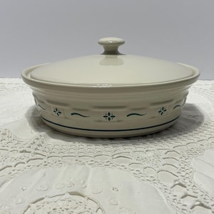 May include: A round, off-white ceramic casserole dish with a matching lid. The exterior features a molded basket-weave texture around the sides, accented by small, dark green floral motifs and thin, horizontal green lines encircling the base.