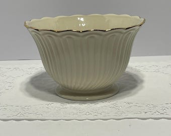 May include: Cream-colored, fluted bowl with a scalloped rim and a gold trim. The bowl sits on a small, round base. The bowl's exterior features vertical, ridged detailing. A decorative piece for the home.