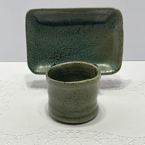 May include: Handmade ceramic set featuring a small, cylindrical cup and a rectangular tray. The pieces are glazed in a mottled green hue, with visible texture on the surfaces. The cup has a slightly rough, organic feel, and the tray has rounded corners.