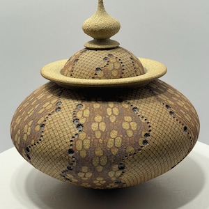 May include: A handcrafted ceramic jar with a lid. The jar is a warm brown color with a textured, patterned design. The lid has a flat, circular base and a decorative finial. The jar is likely used for storage or display.