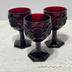 May include: Three deep red glass goblets featuring a textured geometric pattern on the bowl. Each goblet has a thick stem and a hexagonal base. The glass has a translucent quality, showing a repeating grid and circular design on the exterior surface.