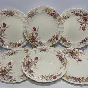 May include: Six off-white ceramic plates with a spiral design and scalloped edges. Each plate features a floral pattern in shades of red, purple, and yellow. The plates are arranged on a white surface.