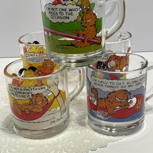 May include: Set of clear glass Garfield mugs. Each mug features a different Garfield cartoon image with text. One mug says "I'm not one who rises to the occasion." Another says "It's not a pretty life but somebody has to live it."