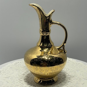 May include: A gold-colored decorative pitcher with a handle and a spout. The pitcher has a bulbous body and a floral pattern. The handle and spout are a lighter gold color. The pitcher is on a white lace doily.