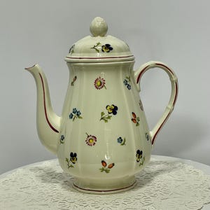 May include: Cream-colored ceramic teapot with a lid and handle, adorned with small, colorful floral patterns. The teapot features a curved spout and a red trim along the edges. The handle has a bamboo-like design.