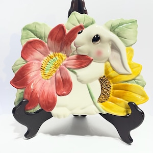 May include: A ceramic bunny figurine with a red flower and yellow sunflower accents. The bunny is white with pink cheeks and is holding the red flower. The figurine is on a black stand, perfect for Easter decor.