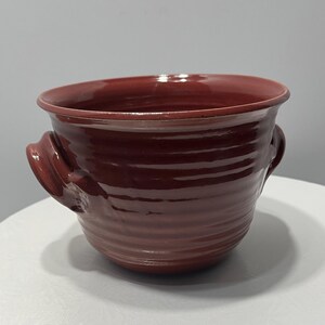 May include: A maroon ceramic bowl with two curved handles. The bowl has a wide, open top and a ridged exterior. The handles are a darker shade of maroon. The bowl is sitting on a white surface.