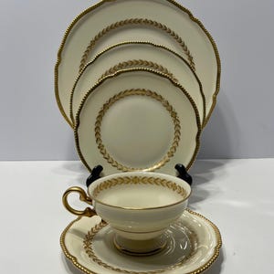 May include: Cream-colored fine china set with gold trim. The set includes a teacup and saucer, plus three plates of varying sizes. Each piece features a gold leaf pattern around the rim.