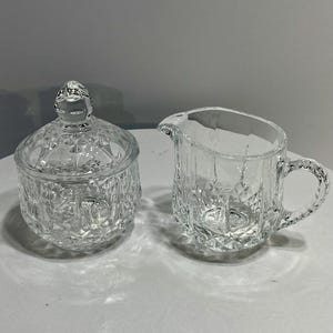 May include: A clear glass sugar bowl with a lid and a creamer pitcher. Both pieces have a diamond-cut pattern. The sugar bowl is round with a knob on top. The creamer has a handle and a spout.