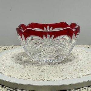 May include: A clear and ruby red cut glass bowl featuring a scalloped rim and intricate diamond-patterned facets. The bowl displays a deep red band along the upper edge with leaf-like motifs carved into the glass, showcasing a reflective finish.