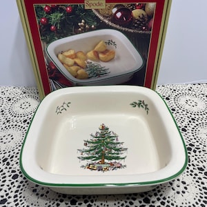 May include: A square, cream-colored ceramic baking dish with a green rim. The interior features a central illustration of a decorated Christmas tree surrounded by gifts, with small sprigs of holly and berries painted on the inner side walls.