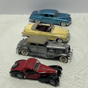 May include: A collection of four vintage-style die-cast model cars arranged in a row. The vehicles include a metallic blue sedan, a pale yellow convertible with a maroon interior, a silver sedan with a tan roof, and a red and black coupe.