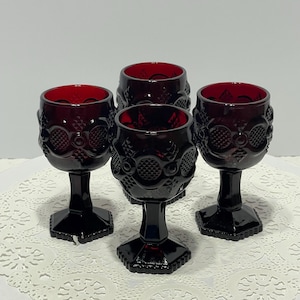 May include: A set of four deep red glass goblets featuring a textured geometric pattern on the bowl and a hexagonal base. Each goblet has a short stem and a rounded cup with a repeating embossed design of circles and cross-hatched squares.