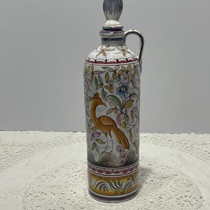 May include: A tall ceramic bottle with a matching stopper and a side handle. The white surface features hand-painted illustrations of a golden-brown bird, blue flowers, and green foliage. The base is decorated with yellow and purple scroll patterns.