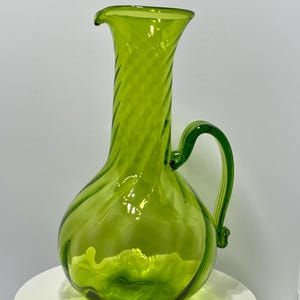 May include: A vibrant green glass pitcher with a handle and a spout. The pitcher has a bulbous base and a tall, slender neck with a spiral design. The pitcher is approximately 12 inches tall.