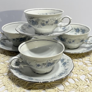 May include: A set of four white teacups and saucers. Each cup and saucer features a delicate blue floral pattern and a silver trim. The cups have curved handles. The saucers are round. The set is arranged on a cream-colored, open-weave surface.