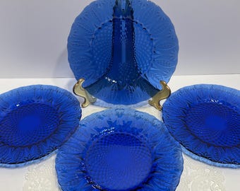 May include: A set of four cobalt blue glass plates. The plates have a textured, floral design. One plate is a serving dish with a raised center, and the other three are round plates. The plates are displayed on a white surface.