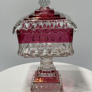May include: A clear and ruby red glass candy dish with a lid. The dish has a square base and a pedestal. The lid is topped with a clear, faceted finial. The dish is likely vintage and suitable for decorative use.