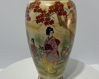 May include: A decorative vase with a cream-colored base and a gold rim. The vase features a painted scene of women in traditional Japanese attire, with red foliage and a gold tree branch.