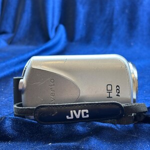 May include: Silver JVC Everio HD camcorder with a black wrist strap. The camcorder has the word "Everio" printed on the top and "HD" printed on the side. The wrist strap has the word "JVC" printed on it.