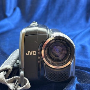 以下が含まれることがあります： 黒いJVCビデオカメラ、Konica Minoltaレンズ付き。ビデオカメラはシルバーのレンズリングとグレーのストラップが付いています。レンズには「35x Optical Zoom/AF」の文字があります。ビデオカメラは青い表面にあります。