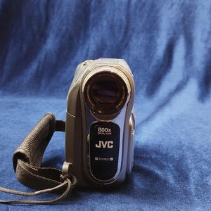 Peut inclure: Caméscope JVC argenté et noir avec une sangle noire. Le caméscope a un objectif et le texte "800x DIGITAL ZOOM" et "JVC" imprimé sur le devant. Le caméscope est sur un fond bleu.
