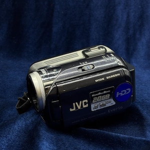 May include: A black JVC Everio camcorder with a silver lens and a blue accent. The camcorder has the text "WIDE SCREEN" and "HARD DISK DRIVE 20GB" printed on it. The camcorder is on a dark blue background.