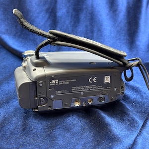 Peut inclure: Caméscope numérique JVC gris, modèle GR-D720E, avec une dragonne noire. Le caméscope porte un marquage CE et d'autres étiquettes. L'appareil est posé sur un tissu bleu.