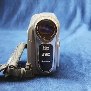 Peut inclure: Caméscope JVC argenté et gris avec une sangle noire. L'appareil photo porte l'inscription "JVC VIDEO LENS" et "800x DIGITAL ZOOM". Le caméscope porte également l'inscription "STEREO".