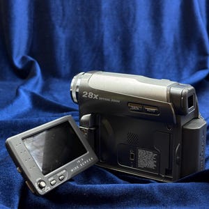 Peut inclure: Caméscope numérique argenté et noir avec écran rabattable. Le caméscope dispose d'un zoom optique 28X et d'un écran large 16:9. L'écran affiche les options de menu et les boutons de commande. Le caméscope est posé sur un fond bleu.