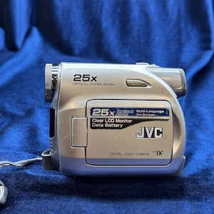 May include: Silver JVC digital video camera with 25x optical hyper zoom, clear LCD monitor, and data battery features. The camera has an auto setting and is labeled "DIGITAL VIDEO CAMERA". The camera is on a blue surface.