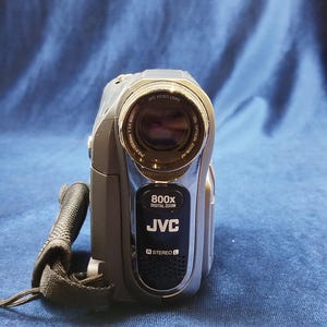 Peut inclure: Caméscope numérique JVC gris avec une sangle noire. L'appareil photo a un objectif argenté et le texte "800x DIGITAL ZOOM JVC STEREO" est visible à l'avant. L'objectif porte l'inscription "JVC VIDEO LENS" et "25x OPTICAL ZOOM/AF".