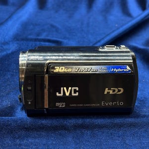 以下が含まれることがあります： 30GBのストレージと32倍光学ズームを備えた、黒いJVC Everioハードディスクカムコーダー。4つの録画モードとハイブリッドテクノロジーを搭載。カムコーダーには「Everio」と「JVC」の文字が印刷されています。