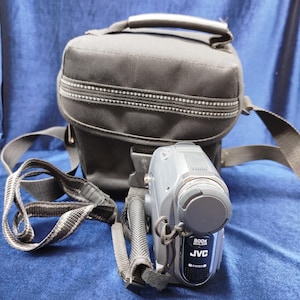 Peut inclure: Caméscope JVC gris avec un étui de transport noir. Le caméscope a une sangle noire et le texte "800x" et "JVC" à l'avant. L'étui noir a une fermeture éclair et une poignée.