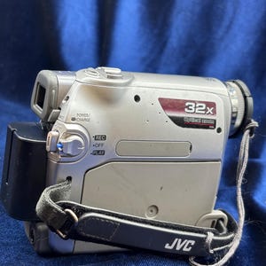 以下が含まれることがあります： 黒いハンドストラップが付いたシルバーのJVCビデオカメラ。ビデオカメラは32倍光学ズームと電源/充電ボタンを備えています。ストラップにはJVCのロゴが入っています。ビデオカメラは青い背景にあります。