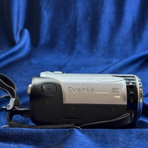 以下が含まれることがあります： 黒とシルバーのEverioビデオカメラ。黒いストラップが付いています。ビデオカメラの側面には「Everio」の文字が印刷されています。ビデオカメラは青色の背景にあります。