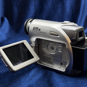 May include: Silver digital camcorder with a flip-out screen. The camcorder has a black lens and the text "25X OPTICAL HYPER ZOOM". The screen is black and the body is silver.