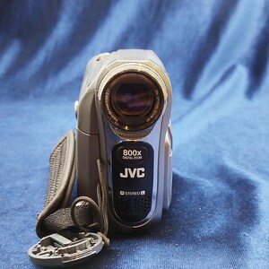 Peut inclure: Caméscope JVC argenté et gris avec un objectif noir et une sangle noire. Le caméscope porte l'inscription "800x DIGITAL ZOOM" et "JVC" sur le devant. Le bouchon de l'objectif est détaché et posé devant le caméscope.