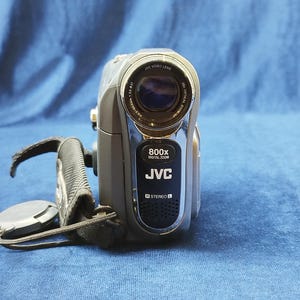 Peut inclure: Caméscope numérique JVC argenté et gris avec une dragonne et un bouchon d'objectif noirs. L'appareil photo porte l'inscription "800x DIGITAL ZOOM" et "JVC" sur le devant. L'objectif est étiqueté "JVC VIDEO LENS".