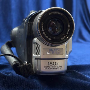 以下が含まれることがあります： シルバーと黒のJVCビデオカメラ、ズームレンズ付き。「JVC VIDEO LENS」と「10X OPTICAL ZOOM/AF」の文字がレンズにあります。ビデオカメラには「150x DIGITAL HYPER ZOOM QUICK RESPONSE AF」の文字があります。