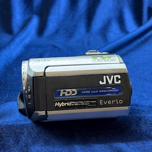 May include: A silver and black JVC Everio camcorder with 34x optical zoom. The camcorder has a Hybrid HDD and SD card recording capability. The device is on a blue surface.