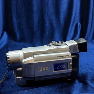 以下が含まれることがあります： 150倍ズームレンズを搭載した、シルバーとブラックのJVCデジタルビデオカメラ。「DIGITAL NIGHT-SCOPE」と「DIGITAL VIDEO CAMERA」の文字が印刷されています。カメラは青い面に置かれています。